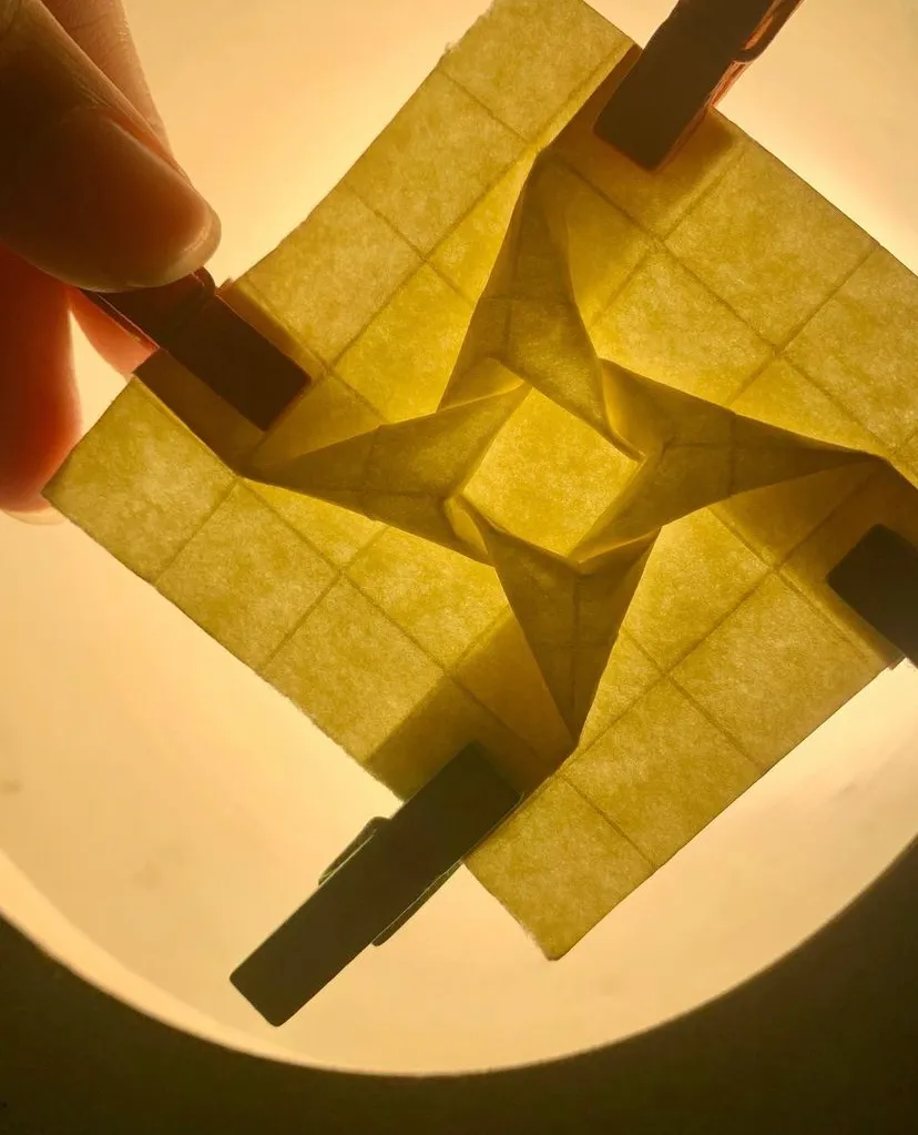 Backlit origami sheet mid-fold, held with wooden pegs